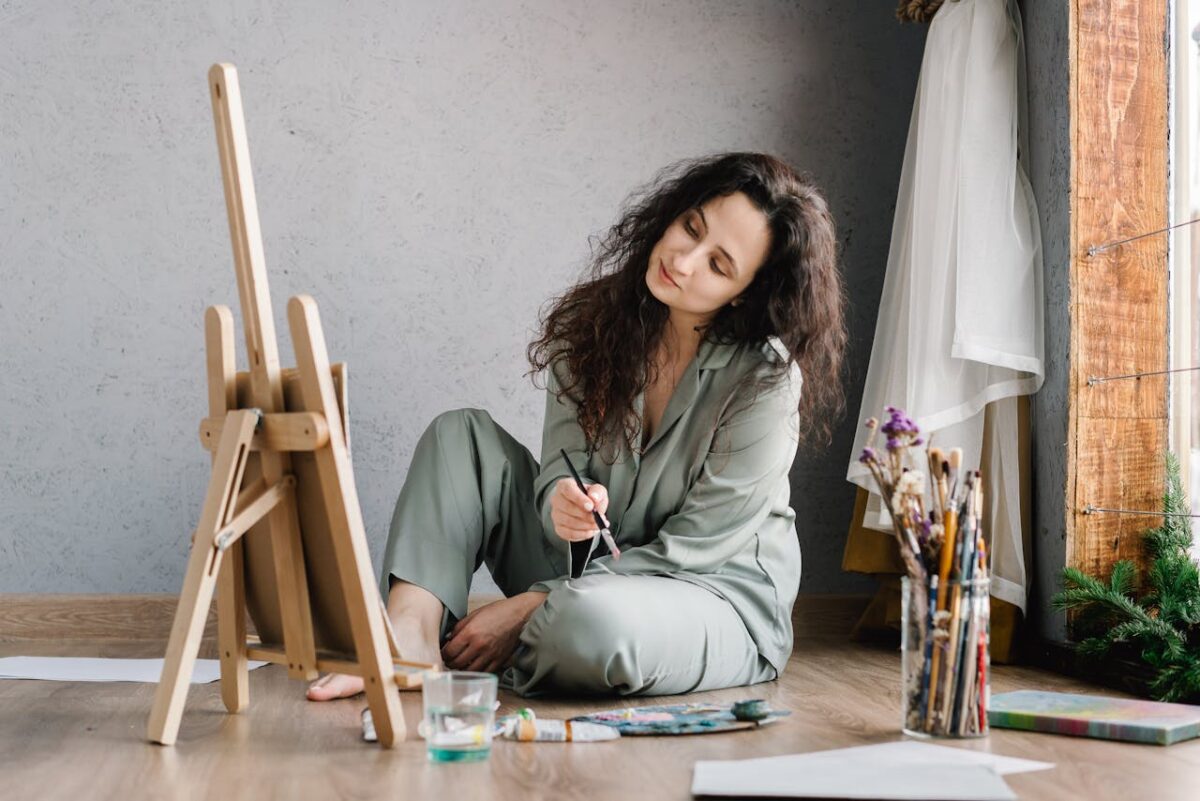 our-mission A woman in comfortable attire paints leisurely by a window with art supplies around her.